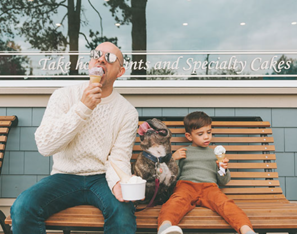 A man, a dog, and a child sit on a bench enjoying ice cream in front of a pastry shop, amidst the charm of luxury 2-bedroom apartments in Lexington Park, MD. | Spark Living